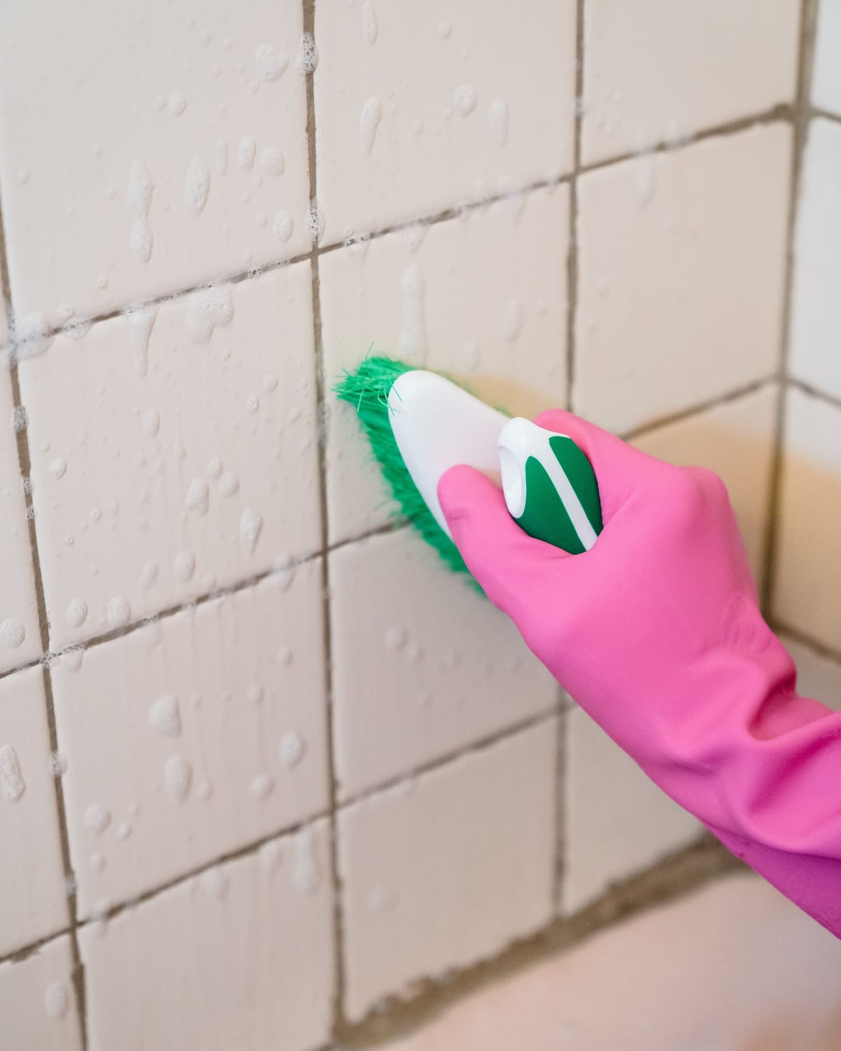 A gloved hand scrubbing the grout lines between white bathroom tiles