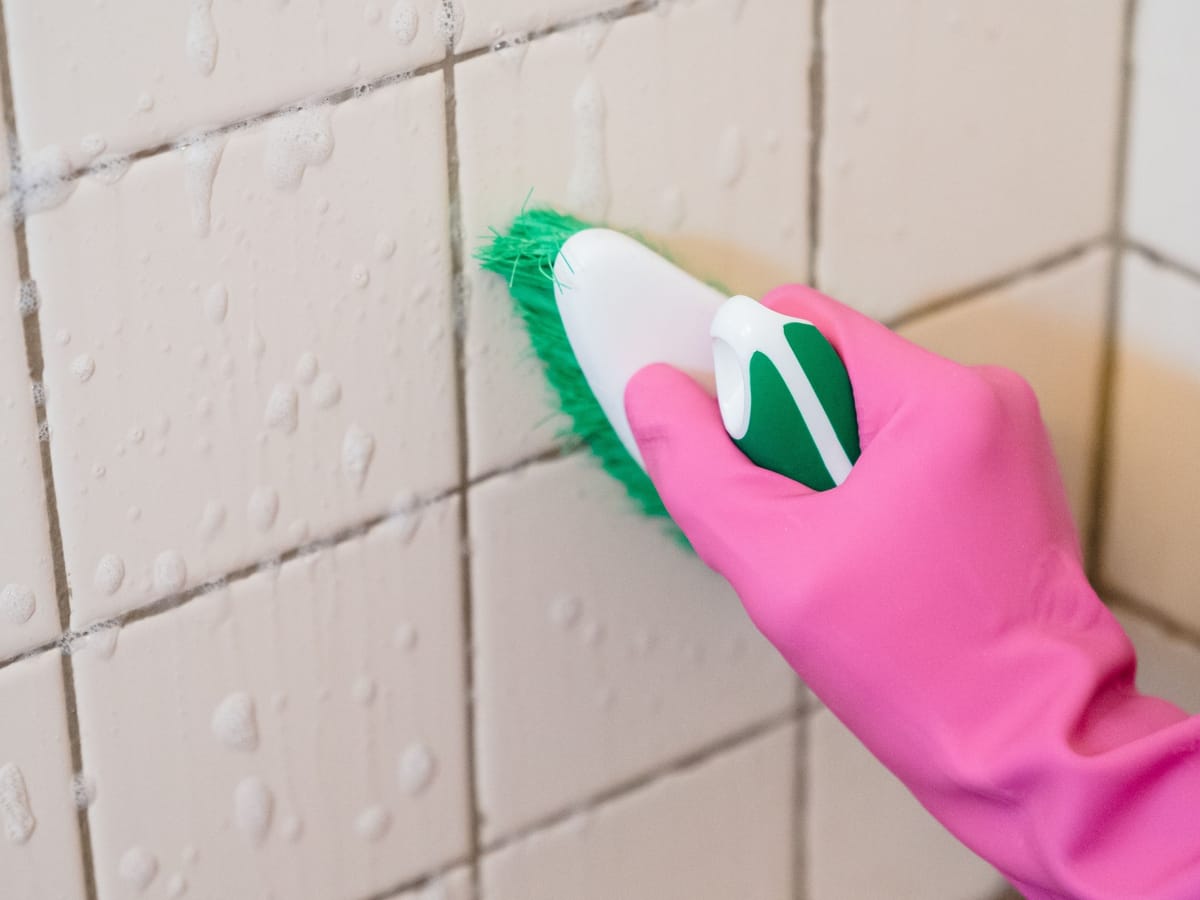 A gloved hand scrubbing the grout lines between white bathroom tiles